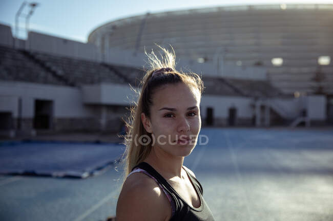 Retrato de una atleta blanca segura de sí misma con el pelo largo y rubio practicando en un estadio deportivo, girando y mirando a la cámara - foto de stock