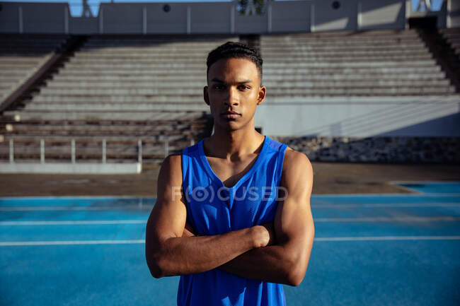 Vista frontal de un atleta masculino de raza mixta confiado con un chaleco azul practicando en un estadio deportivo, de pie con los brazos cruzados mirando directamente a la cámara - foto de stock