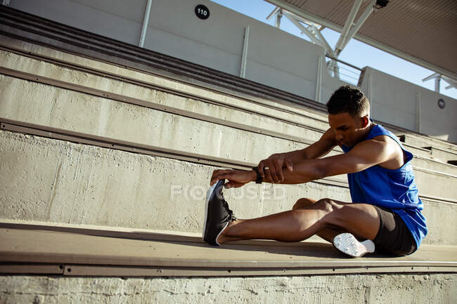 Vue latérale d'un athlète masculin de race mixte pratiquant dans un stade de sport, assis dans les gradins et s'étirant, tirant sur son pied — Photo de stock
