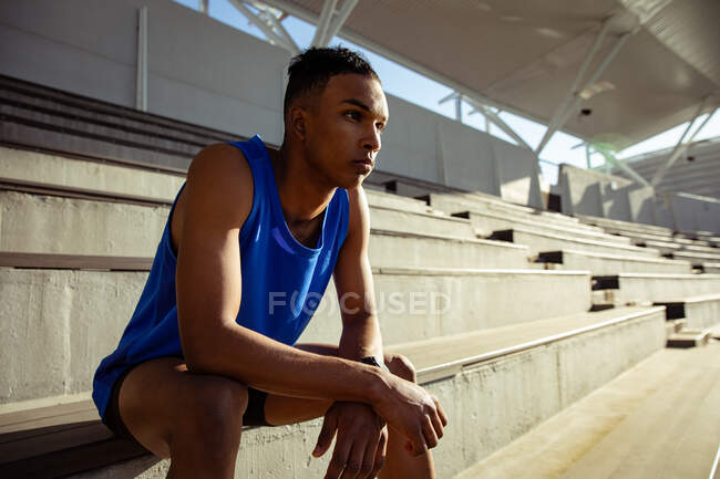 Vista lateral de un atleta masculino de raza mixta practicando en un estadio deportivo, sentado en las gradas y enfocado antes de una carrera - foto de stock