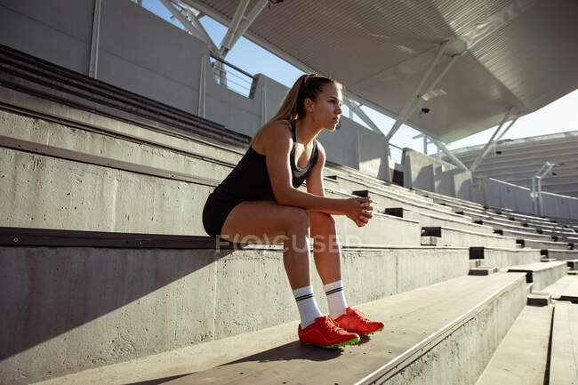 Vista lateral de una atleta caucásica practicando en un estadio deportivo, sentada en las gradas y enfocada antes de una carrera - foto de stock