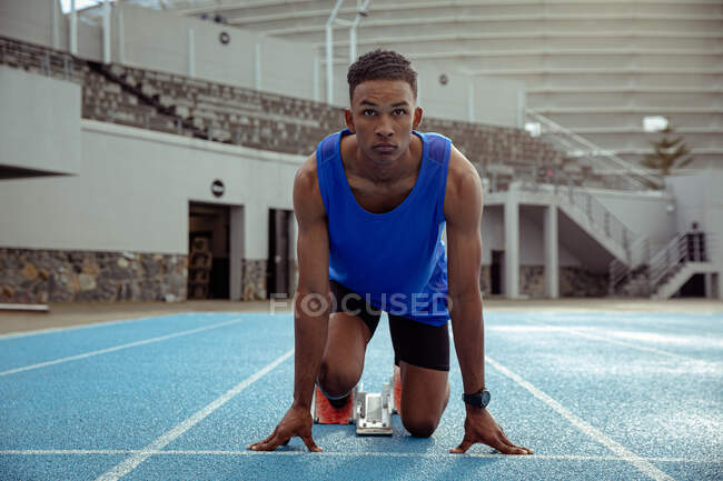 Front view of a mixed race male athlete practicing at a sports stadium, in position on the starting blocks, preparing to sprint, with his head up — Stock Photo