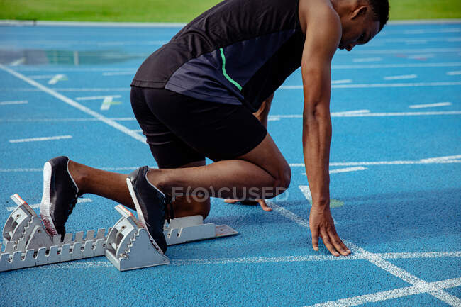 Vista lateral de un atleta masculino de raza mixta practicando en un estadio deportivo, en posición sobre bloques de salida, preparándose para correr - foto de stock