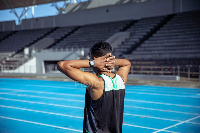 Vista lateral de un atleta masculino de raza mixta practicando en un estadio deportivo, sosteniendo sus manos sobre su cuello, enfocándose antes de una carrera - foto de stock