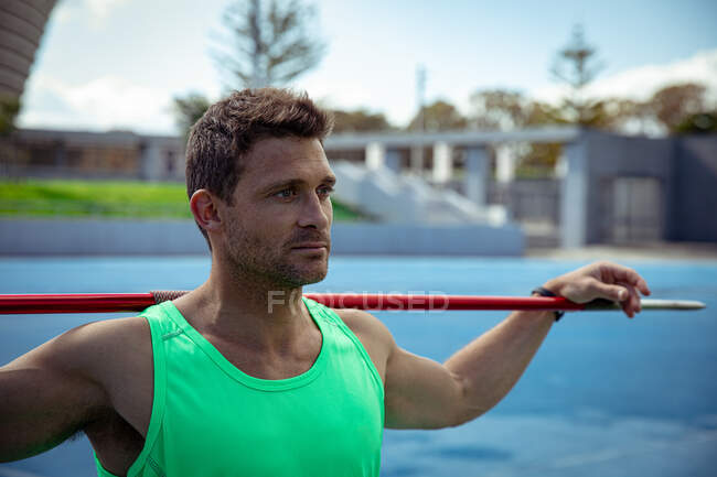 Front view of a Caucasian male athlete practicing at a sports stadium, resting his hands on a javelin across his shoulders and looking away — Stock Photo