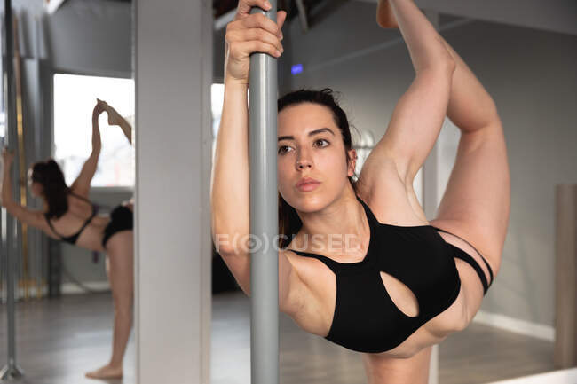 Передній вигляд принадної привабливої кавказької жінки, яка насолоджується поул-танцями в студії, тримаючи стовп однією рукою і ногу з іншого боку, розтягуючи. — стокове фото