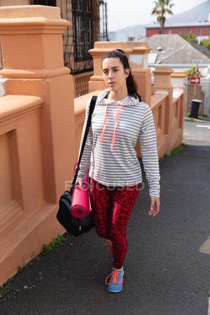 Vorderansicht einer fitten Kaukasierin auf dem Weg zum Fitnesstraining an einem bewölkten Tag mit Sporttasche und Yogamatte — Stockfoto