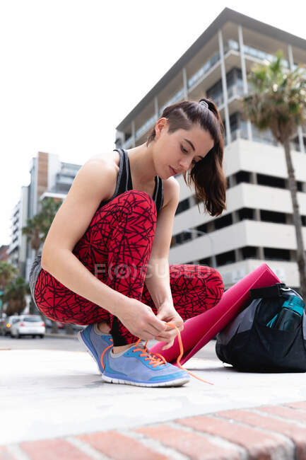 Вид спереди на белокурую женщину, идущую на тренировку в облачный день, несущую спортивную сумку и коврик для йоги, стоящую на коленях на улице и завязывающую шнурки — стоковое фото