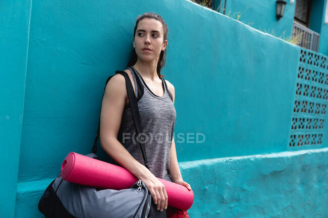 Vista frontal de una mujer caucásica en forma en su camino al entrenamiento de fitness en un día nublado, llevando una bolsa de deporte y una esterilla de yoga, apoyada en una pared azul - foto de stock