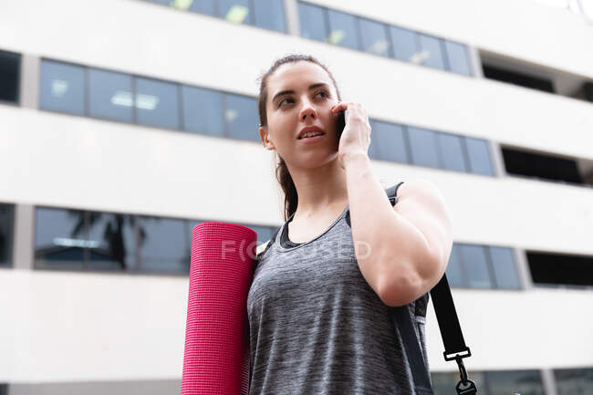 Vista frontal de una mujer caucásica en forma en su camino al entrenamiento de fitness en un día nublado, llevando una bolsa de deporte y una esterilla de yoga, hablando en un teléfono inteligente y caminando por la calle - foto de stock