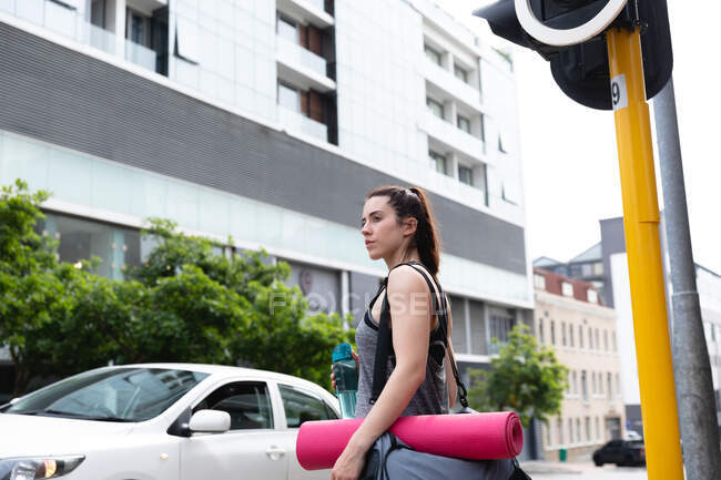 Vista lateral de uma mulher caucasiana em forma em seu caminho para o treinamento de fitness em um dia nublado, esperando para atravessar uma estrada, carregando um saco esportivo e um tapete de ioga, segurando uma garrafa de água — Fotografia de Stock