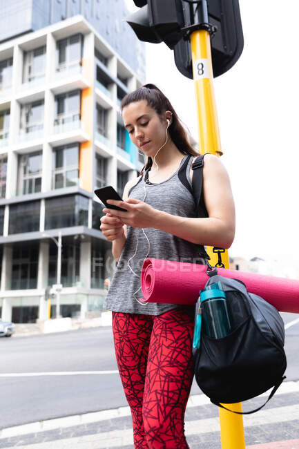 Front view of a fit Caucasian woman on her way to fitness training on a cloudy day, standing in the street carrying a sports bag and a yoga mat, using her smartphone and wearing earphones — Stock Photo