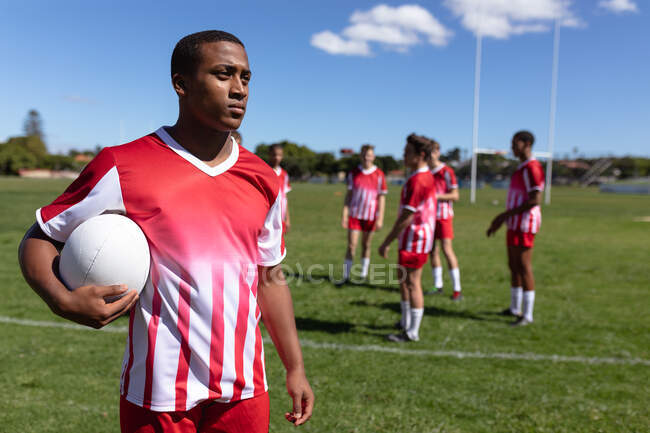 Front view of a teenage mixed race male rugby player wearing red and white team strip, standing on a playing field, holding a rugby ball with other players warming up in the background — Stock Photo