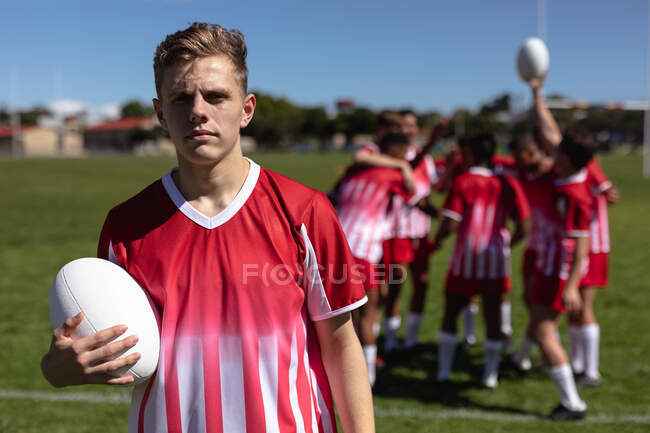 Front view of a teenage Caucasian male rugby player wearing red and white team strip, standing on a playing field, holding a rugby ball and looking at camera, with other players warming up in the background — Stock Photo