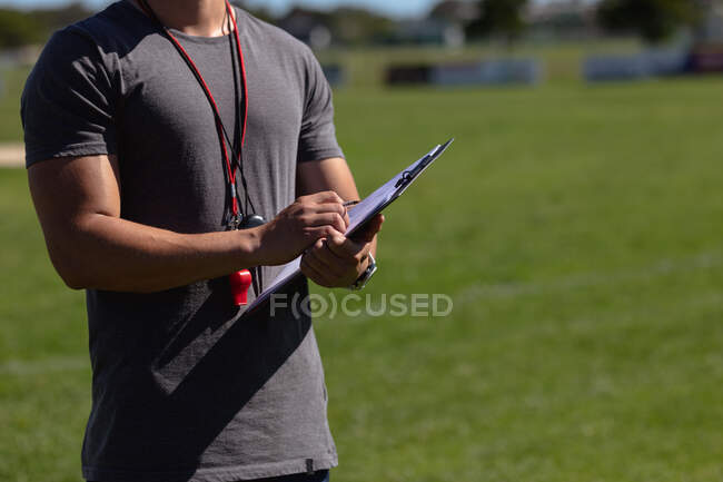 Vista frontal sección central del entrenador de rugby masculino de pie en el campo de juego haciendo notas en su portapapeles - foto de stock