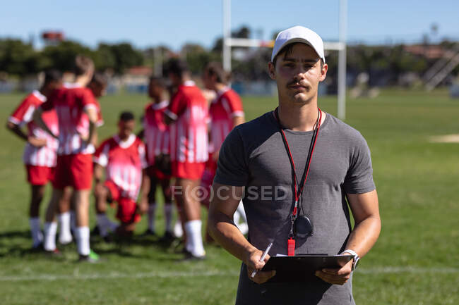 Front view of a Caucasian male rugby coach standing on a playing field holding a clipboard and looking to camera, with a teenage multi-ethnic male team of rugby players wearing their team strip standing in the background — Stock Photo