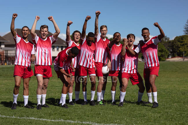 Vista frontal de un grupo de jugadores de rugby masculinos multiétnicos adolescentes que usan tira de equipo roja y blanca, celebrando una victoria, de pie con los brazos levantados en un campo de juego y mirando a la cámara animando - foto de stock