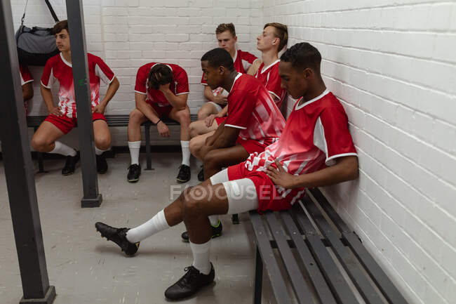 Вид сбоку на группу подростков многонациональных игроков в регби, одетых в красно-белую командную одежду, сидящих и отдыхающих в раздевалке после игры в регби — стоковое фото