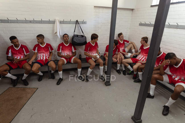 Вид спереду групи підлітків-мультиетнічних чоловіків регбістів в червоно-білій командній смузі, сидячи і відпочиваючи в роздягальні після гри в матчі — стокове фото