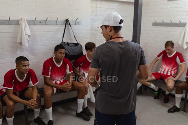 Rear view of a Caucasian male rugby coach standing in the changing room talking to a teenage multi-ethnic male team of rugby players wearing their team strip, sitting and listening — Stock Photo