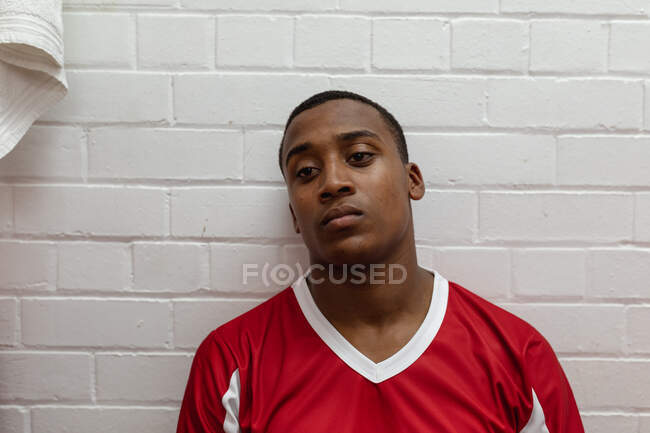 Portrait close up of a teenage mixed race male rugby player wearing red and white team strip, sitting and resting in the changing room after a match — Stock Photo