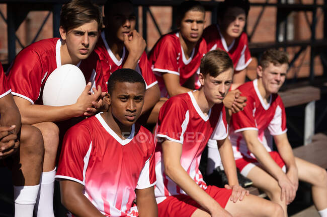 Frontansicht einer Gruppe multiethnischer männlicher Rugby-Spieler, die rot-weiße Mannschaftskleidung tragen und sich ein Spiel von der Tribüne aus ansehen — Stockfoto