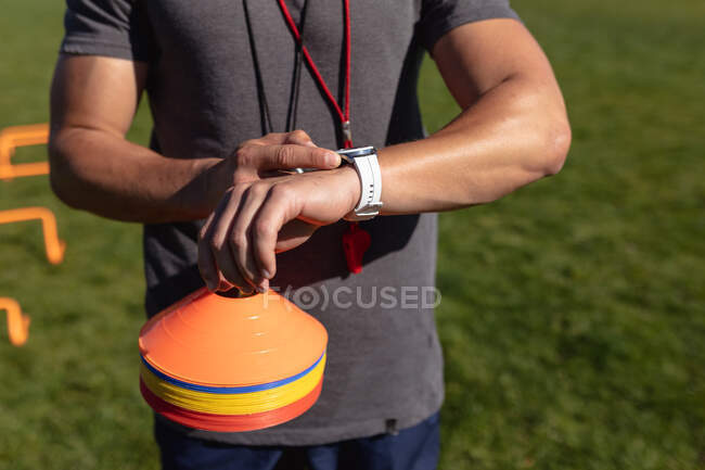 Vorderansicht Mittelteil eines männlichen Rugby-Trainers, der auf dem Spielfeld steht und auf seine Uhr blickt und einen Stapel Plastikmarker in der Hand hält — Stockfoto