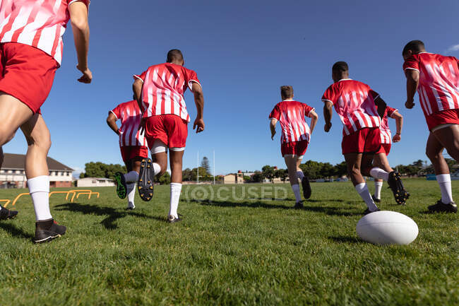 Niedrige Rückansicht einer multiethnischen Männermannschaft mit Rugbyspielern, die ihren Mannschaftsstreifen tragen, gemeinsam auf dem Spielfeld laufen, blauer Himmel im Hintergrund — Stockfoto