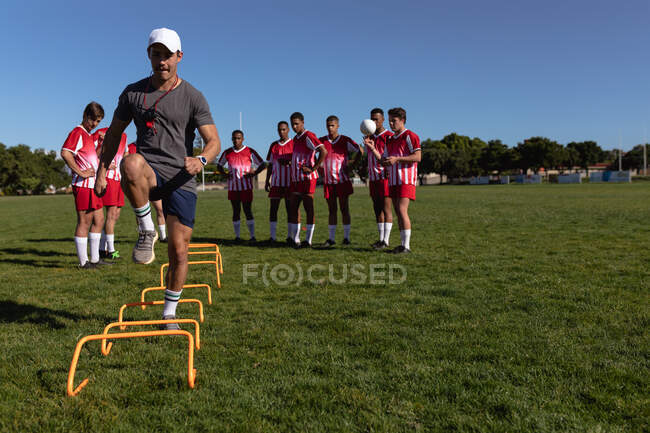 Vista frontal de un entrenador de rugby masculino caucásico mostrando un ejercicio a un equipo masculino multiétnico adolescente de jugadores de rugby usando su tira de equipo, de pie detrás de él en el campo de juego - foto de stock