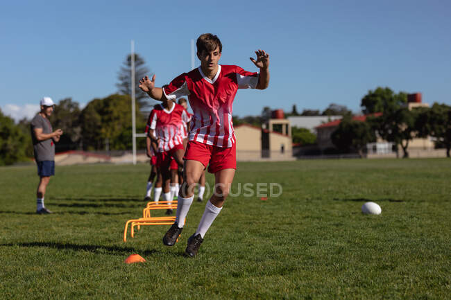 Vue de face d'un joueur de rugby adolescent caucasien s'exerçant pendant une séance d'entraînement sur le terrain de jeu, franchissant de bas obstacles, avec son entraîneur et d'autres joueurs en arrière-plan — Photo de stock