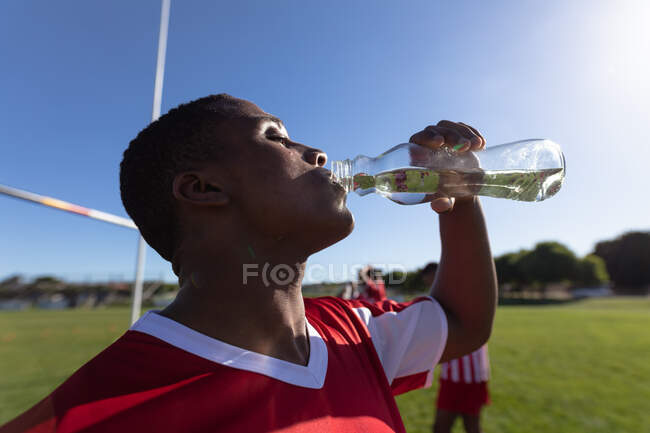 Vista lateral de perto de um jogador de rugby mestiço adolescente vestindo faixa de equipe vermelha e branca, em pé em um campo de jogo, água potável, com os outros jogadores por trás. — Fotografia de Stock