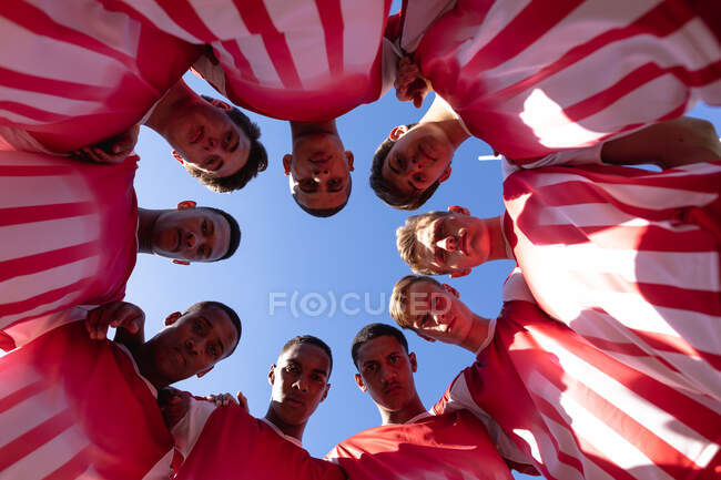 Vista ad angolo basso guardando una squadra maschile multietnica adolescente di giocatori di rugby che indossano la loro striscia di squadra, abbracciandosi e guardando la telecamera contro un cielo blu — Foto stock