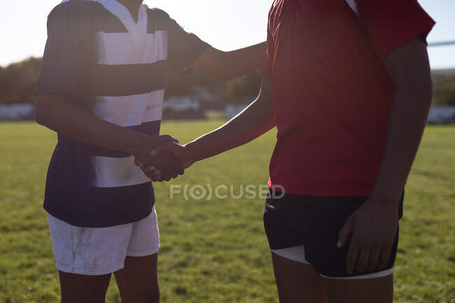 Vista lateral na metade da seção de dois jogadores de rugby masculinos de equipes adversárias usando sua tira de equipe, apertando as mãos em pé no campo de jogo antes de uma partida — Fotografia de Stock