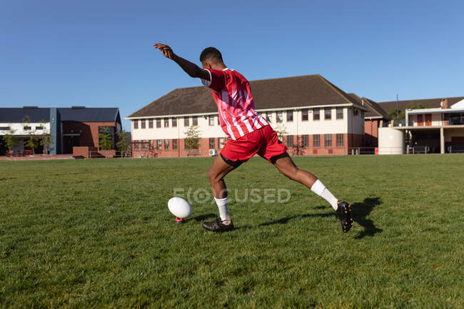 Вид сбоку на юношеского регбиста смешанной расы, одетого в красно-белую команду, пинающего регбийный мяч на игровом поле, со школьным зданием на заднем плане — стоковое фото