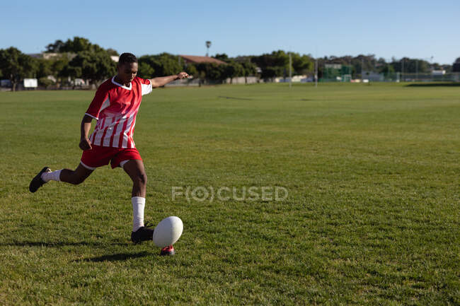 Vista frontal de um jogador de rugby mestiço adolescente vestindo faixa vermelha e branca, chutando uma bola de rugby em um campo de jogo. — Fotografia de Stock