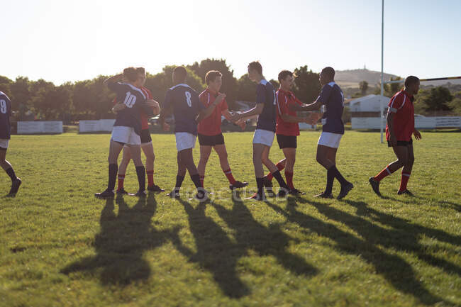 Vista lateral de dos equipos masculinos multiétnicos adolescentes de jugadores de rugby que usan su tira de equipo, saludándose en el campo de juego, dándose la mano antes del inicio de un partido - foto de stock