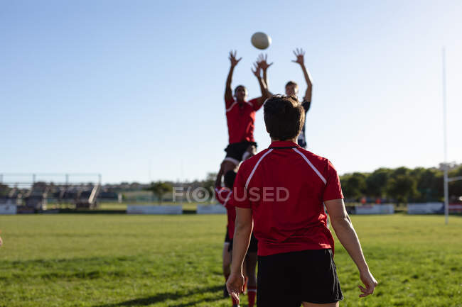 Rückansicht eines jugendlichen kaukasischen männlichen Rugbyspielers in roter Mannschaftskleidung, der auf einem Spielfeld steht und zwei andere Spieler beobachtet, die von Teamkollegen mit den Händen in die Luft gehoben werden und nach dem Ball greifen. — Stockfoto