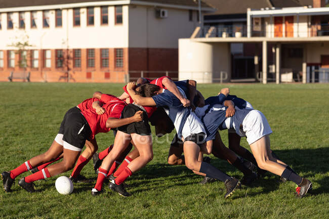 Seitenansicht von zwei multiethnischen Männermannschaften mit Rugby-Spielern, die ihre Mannschaftsstreifen tragen, in einem Gedränge während eines Spiels auf einem Spielfeld mit Gebäuden im Hintergrund — Stockfoto