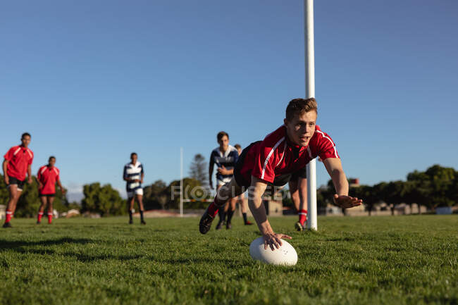 Vista frontal de un jugador de rugby masculino caucásico adolescente con tira roja del equipo, en un campo de juego, buceando con la pelota para anotar un intento durante un partido, con compañeros de equipo y jugadores del equipo contrario en el fondo - foto de stock