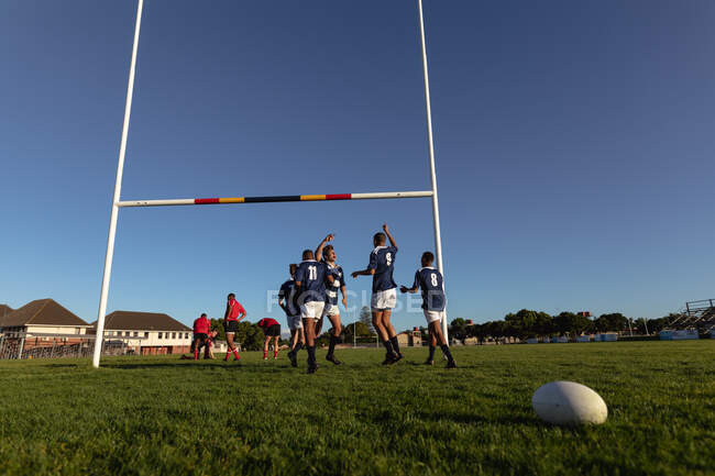 Vue latérale d'un groupe de joueurs de rugby masculins multiethniques adolescents portant une bande bleue et blanche, célébrant la victoire, levant les mains, se tenant sur un terrain de jeu avec d'autres joueurs derrière et une balle de rugby posée sur le terrain. — Photo de stock