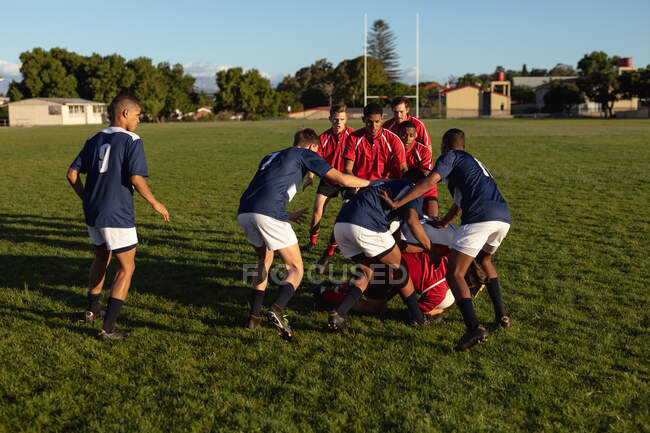 Vista lateral de dos equipos masculinos multiétnicos adolescentes de jugadores de rugby que usan sus tiras de equipo, en acción durante un partido de rugby en un campo de juego. - foto de stock