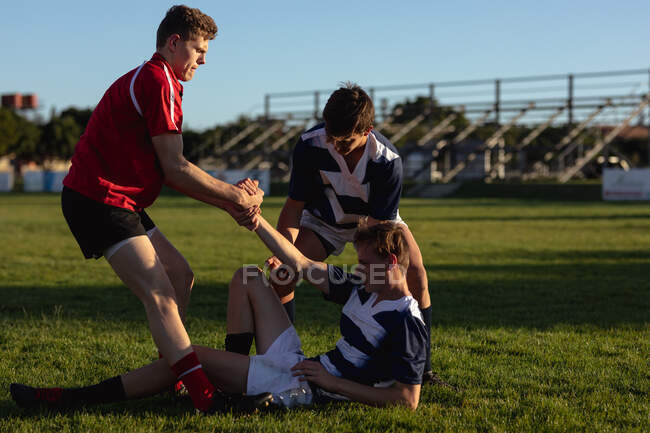 Vista lateral de un jugador de rugby adolescente caucásico ayudando a otro jugador de rugby adolescente caucásico del otro equipo desde el suelo con la ayuda de su compañero de equipo durante un partido de rugby en un campo de juego - foto de stock