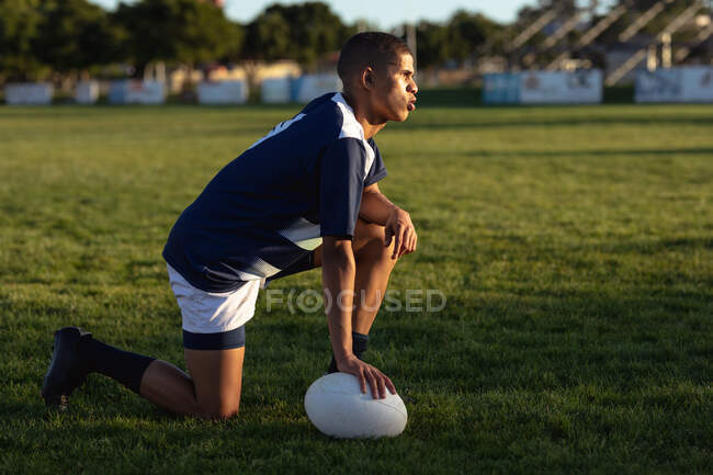 Side view of a teenage mixed-race male rugby player wearing blue and white team strip, preparing to kick the rugby ball, kneeling down on a playing field. — Stock Photo
