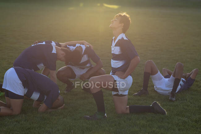 Vista laterale dei giocatori di rugby maschi multietnici adolescenti che indossano strisce di squadra blu e bianche, che riposano su un campo di gioco dopo una partita — Foto stock
