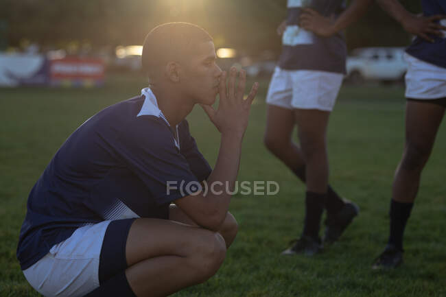 Vista lateral de un jugador de rugby masculino adolescente de raza mixta que usa tira de equipo azul y blanco, se agacha en un campo de juego, descansa después de un partido de rugby, con otros jugadores detrás, retroiluminado - foto de stock