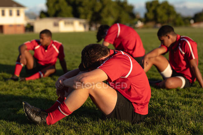 Вид збоку чотирьох підлітків багатоетнічних чоловіків регбі в червоній командній смузі, сидячи і відпочиваючи на сонці на ігровому полі після матчу регбі — стокове фото