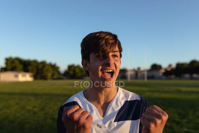 Ritratto ravvicinato di un adolescente bianco giocatore di rugby maschile che indossa una striscia di squadra blu e bianca, in piedi su un campo di gioco e tifo, urlando e alzando le mani al sole durante una partita di rugby — Foto stock