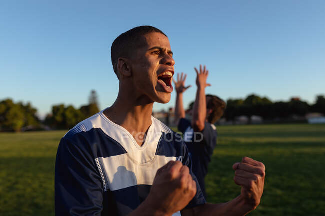 Вид спереди на юношеского регбиста смешанной расы в сине-белой команде, стоящего на игровом поле и приветствующего, кричащего и поднимающего руки в честь победы, с другим игроком с поднятыми руками на заднем плане — стоковое фото