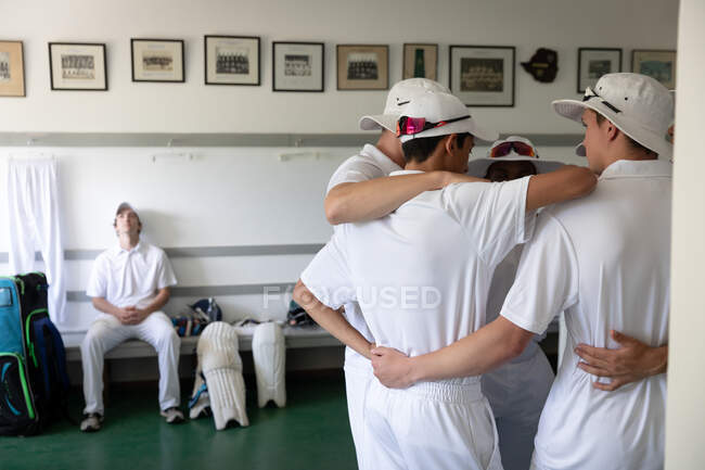 Vista laterale di un gruppo di giocatori di cricket maschi multietnici adolescenti che indossano i bianchi, rannicchiati in uno spogliatoio, con un altro giocatore appoggiato su una panchina. — Foto stock