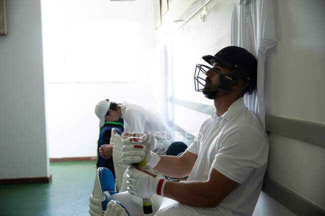 Vista lateral de un jugador de cricket masculino de raza mixta que lleva casco blanco y cricket, sentado en un banco en un vestuario, con otro jugador sentado en el fondo - foto de stock
